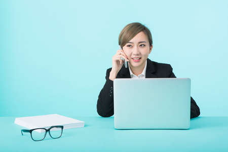 A young woman in a black suit smiling with her smartphone in her ear while operating a computer on a green desk.の写真素材