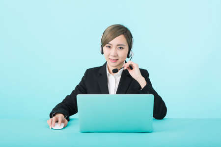 A young woman wearing a headset in front of a green background and wearing a black suit heading to her laptopの写真素材
