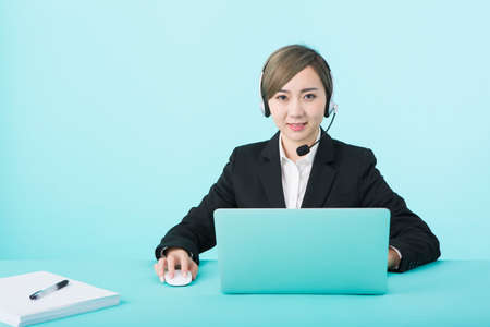 A young woman wearing a headset in front of a green background and wearing a black suit heading to her laptopの写真素材