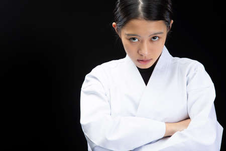 a young woman with dark hair who wears a white dress in front of a black background, tightens her black belt, puts on her arms and looks seriousの写真素材