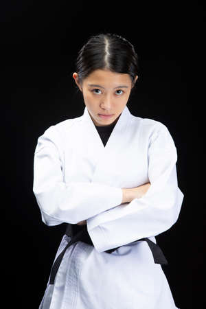 a young woman with dark hair who wears a white dress in front of a black background, tightens her black belt, puts on her arms and looks seriousの写真素材