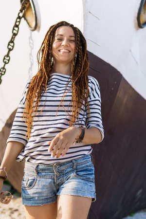 a girl with a dreadlocked hairstyle poses on the beach near a ship in summer, the sea and sand on a...