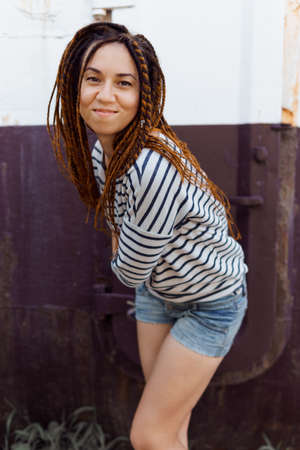 a girl with a dreadlocked hairstyle poses in summer in the shade of trees, branches with green...