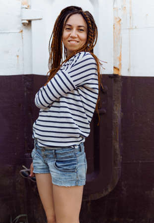 a girl with a dreadlocked hairstyle poses in summer outdoor, bright sunlight, dressed in a plaid shirt and shortsの写真素材