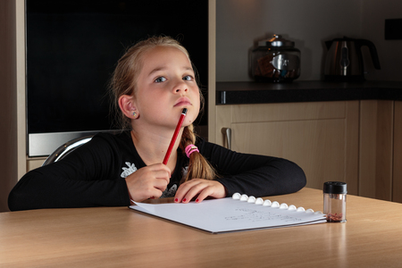 Young girl doing homework at the kitchen table. Thinking and holding pencil against her mouth while writing random wordsの写真素材