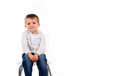 Adorable cute little boy sitting on a stool wearing a white shirt and a blue jeans. Isolated on whiteの写真素材
