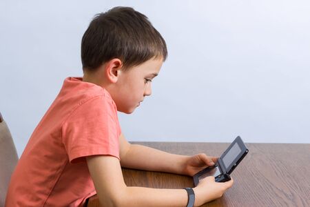Young boy playing handheld video game at a table. Against grey background
