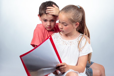 Caucasian schoolchildren studying, a cute girl holding notes, explains a boy who looks confused and strikes his hand in his hair on light background. Education conceptの写真素材
