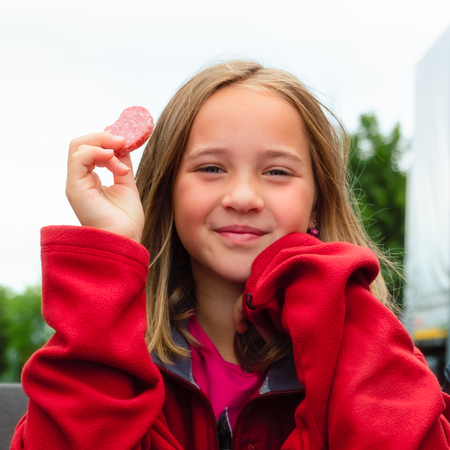 Portrait of a cheerful 10 year old girl with red sweater holding a piece of salami sausage smiling outdoorsの写真素材