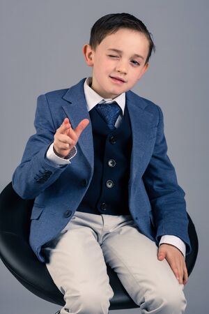 Portrait of handsome 8 year old boy dressed up in suit sitting on a stool winks and points fingers, cool attitudeの写真素材