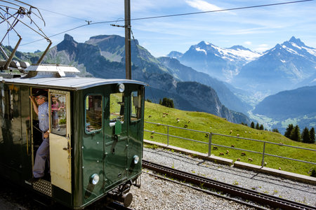 Schynige Platte, Bernese Oberland, Switzerland - August 1 2019: driver standing in doorway of green tourist train on Schynige Platte. Green pasture and high mountain peaks in the backgroundのeditorial素材