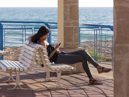 Nahariya, Israel - May 30, 2016: Young girl sits on a sunset on the beach, and wrote a message on a mobile phone in Nahariya, Israelのeditorial素材