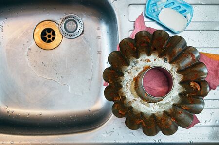Single dirty baking metal form. Leftovers of fresh baking. Placed on top of a steel kitchen sink with water droplets all around. Pink washcloth and slimy white soap bar. No people, real raw shotの写真素材