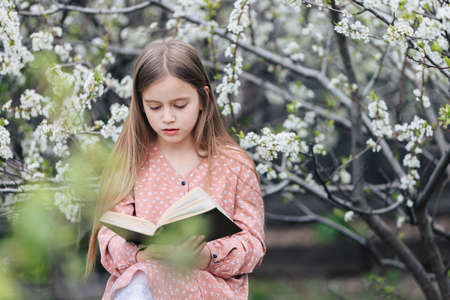 Little girl is reading a book near a flowering tree in the garden.の写真素材
