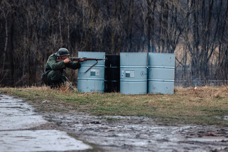 VIKHOREVKA, RUSSIA - May 09, 2021: Reconstruction of the 1941-1945 hostilities between the armies of Germany and the USSR. A German soldier fires a rifle and hides behind barrels.のeditorial素材