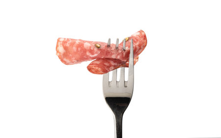 Two thin slices of cooked smoked sausage with green pepper on a fork. Fork with sausage isolated on a white background. Close up. Copy space.の写真素材