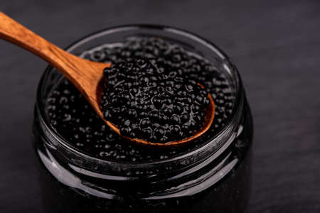 A wooden spoon with black caviar over a glass jar of caviar. Selective focus on black caviar in a wooden spoon.の写真素材