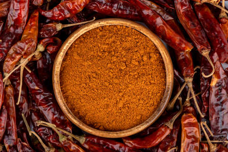 Top view of a wooden bowl with ground red pepper among dry hot pepper pods. close up.