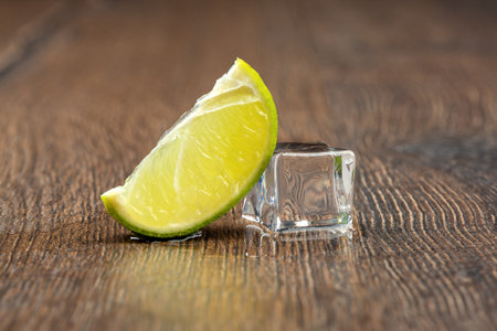 Slice of lime and a melting ice cube with drops of water on the table. Pure ice in the form of a cube. Frozen water. Fake or artificial acrylic or plastic ice cubes. Wooden background.の写真素材