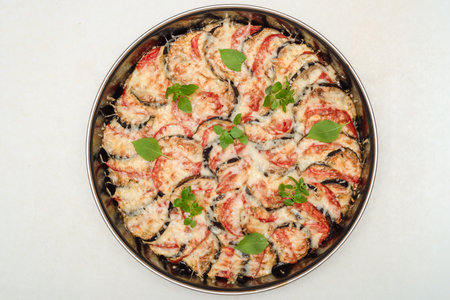 Baking dish with baked eggplants and tomatoes under grated cheese, decorated with fresh basil leaves.の写真素材