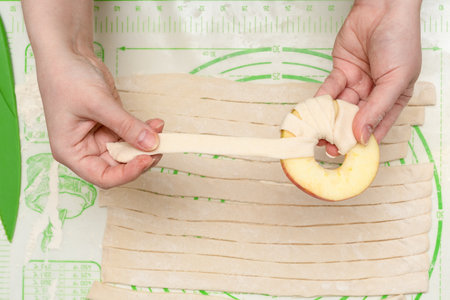 A young woman wraps a strip of puff pastry around an apple circle before baking it in the oven. Home cooking.の写真素材