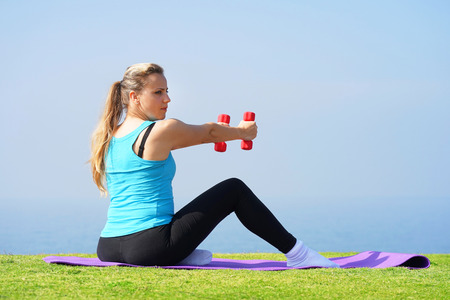 Young girl sitting on a rug on the grass with dumbbells in hands on the background of the sea.の写真素材
