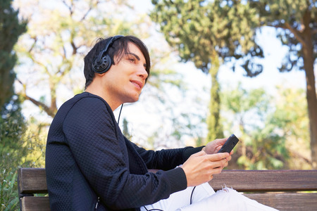 The guy is sitting on the bench, using a smartphone to listen to music on headphones. Joyful attractive man listening to music on headphones while enjoying sitting in the park. Lifestyle conceptの写真素材
