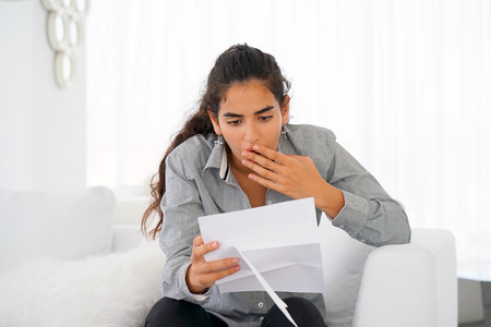 Woman reads negative news in a letter at home on the couch. The shaken business manager of the beauty girl received a notice of dismissal from the company, surprised. An agitated girl without joy.の写真素材