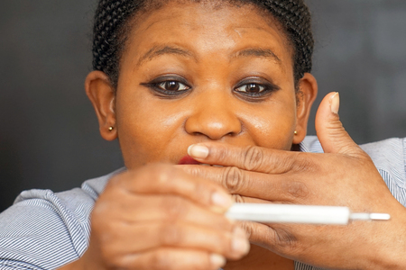 African woman holding thermometer in hands, measuring body temperature while suffering from influenza. Frustrated female with thermometer feeling sick.の写真素材