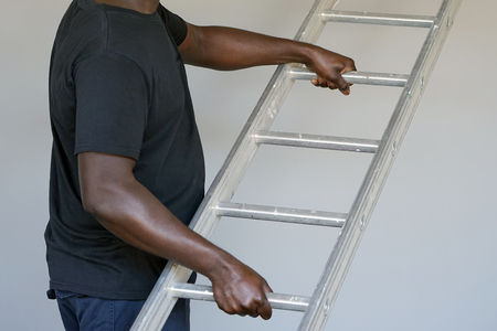 An electrician holds an aluminum ladder. The builder carries an aluminum stepladder on gray background. Worker of the plant with aluminum rung ladderの写真素材