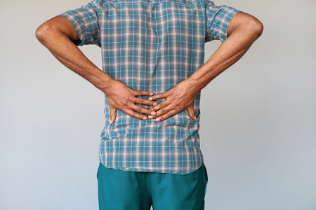 Close-up view of a young man with pain in kidneys on gray background. Young man with back ache clasping her hand to her lower back. Man suffering from ribbing pain, waist pain.の写真素材