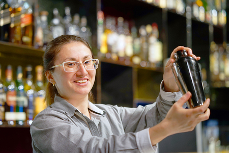 Professional bartender girl holding in hands a shaker with a fresh delicious cocktail. Barmaid shaking a cocktail shaker as she stands behind the bar mixing a drink for a client.の写真素材
