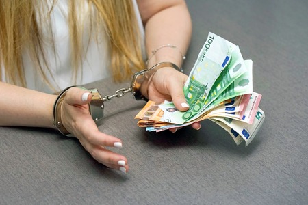 Businesswoman in office in handcuffs holding a bribe of euro banknote. Close-up woman hands in handcuffs. Arrested terrorist. Financial crime, dirty money and corruption concept. Selective focus.の写真素材