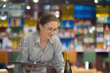 Waitress carries a bottle of white wine with two wine glasses on a tray to the client in the hotel bar. Girl in glasses. The concept of service.の写真素材