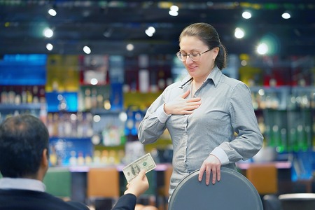 Waitress gets a tip from the client in the bar of the hotel. The girl in the vow. A bartender woman is happy to receive a tip at work. Shelves with bottles of alcohol in the background. Emotions.の写真素材