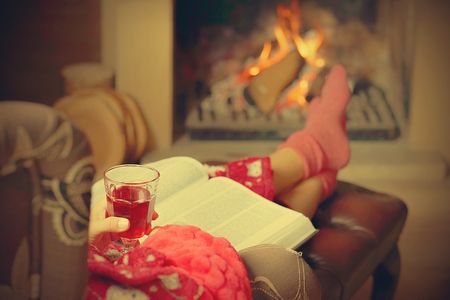 Woman legs with socks sitting near the fireplace with a warm background. Woman in warm socks resting near the fireplace at home with a book and a glass of red wine. Toning.の写真素材