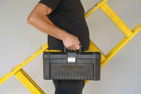 Electrician holds tool box and yellow wooden ladder. The builder carries a wooden ladder and toolbox on a gray background. Breeder with tool box and yellow wooden ladder.の写真素材