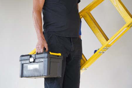 Electrician holds tool box and yellow wooden ladder. The builder carries a wooden ladder and toolbox on a gray background. Breeder with tool box and yellow wooden ladder.の写真素材