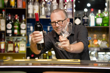 The bartender holds a bottle of red wine and wine glasses in the hotel bar. A bartender man gives a waiter a bottle of wine and glasses. The service concept.の写真素材