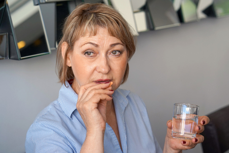 Sick elderly woman holding pill glass of water at home on the sofa. Depressed unhealthy woman, about to take antidepressant pill, emergency contraceptive, painkiller for painful periods.の写真素材