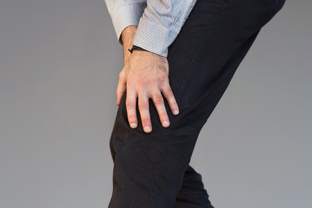 Pain In Knee. Close-up Businessman Leg With Painful Kneeson isolated on gray background. Man Feeling Joint Pain, Having Health Issues And Touching Leg With Hands. Body And Health Care Concept.の写真素材