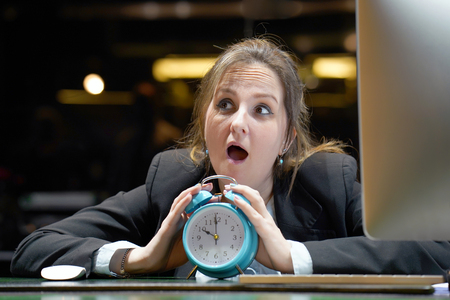 Woman holds an alarm clock in her hand in the workplace. female office worker holds an alarm clock in her hand at her desk. Woman holding a clock. Time management concept.の写真素材