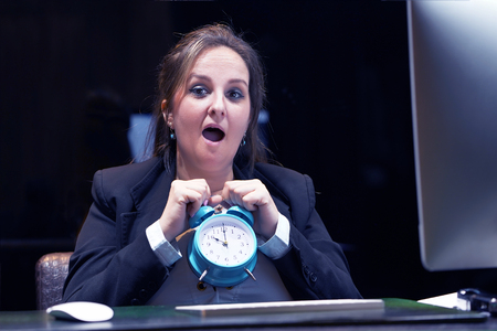 Woman holds an alarm clock in her hand in the workplace. female office worker holds an alarm clock in her hand at her desk. Woman holding a clock. Time management concept.の写真素材