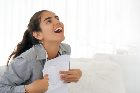 Young woman enjoying good news in writing. The girl reads a letter with good news sitting on the couch. An euphoric girl is happy after reading good news in a written letter, approving a loan.の写真素材