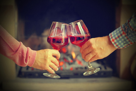 Man and woman hand toasting wine glasses in front of fireplace at home. Elderly couple have romantic dinner with red wine over fireplace background. Romantic winter night for couple in love. Toning.の写真素材