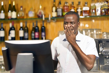 Male bartender made a mistake in the bill, emotion. A barmaid registrating new order by cash register. A restaurant worker registrating new order by cash register. The concept of service.の写真素材