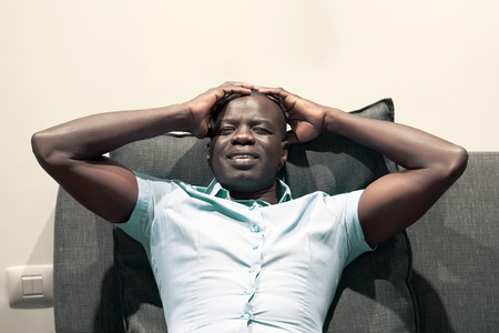 African man suffering from headache migraine pain at home on sofa. Health problem, stress and depression. Male holds head with hand. Concept of health.の写真素材