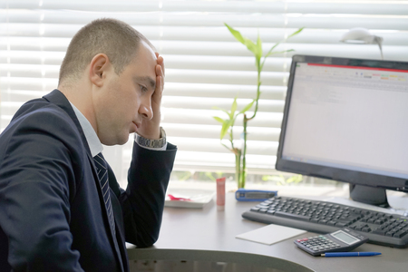 Social worker working at her desk suffering from headache migraine pain. Man hospital secretary sitting at her workplace. Health problem, stress and depression. Male holds head with hand.の写真素材