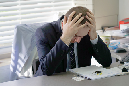 Social worker working at her desk suffering from headache migraine pain. Man hospital secretary sitting at her workplace. Health problem, stress and depression. Male holds head with hand.の写真素材