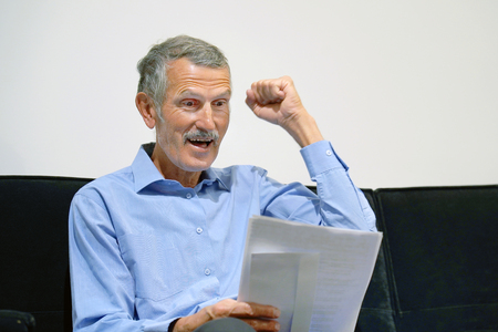 Elderly man enjoying good news in writing. The male reads a letter with good news sitting on the couch. An euphoric male is happy after reading good news in a written letter, approving a loan.の写真素材